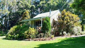 A small, single-story house with a porch, surrounded by lush greenery and trees.