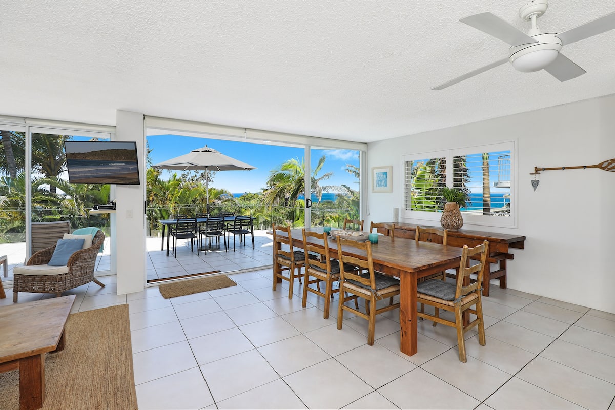 A spacious dining area with a wooden table and chairs, large windows with a view of the ocean, and a ceiling fan.