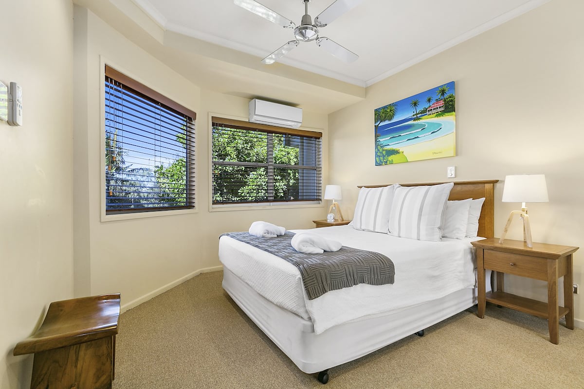 A hotel room with a bed, a nightstand, a ceiling fan, and a painting of a beach scene.