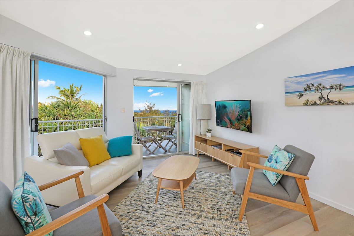 A modern living room with a sofa, armchairs, a coffee table, and a TV. There is a balcony with a view of palm trees and the sky.