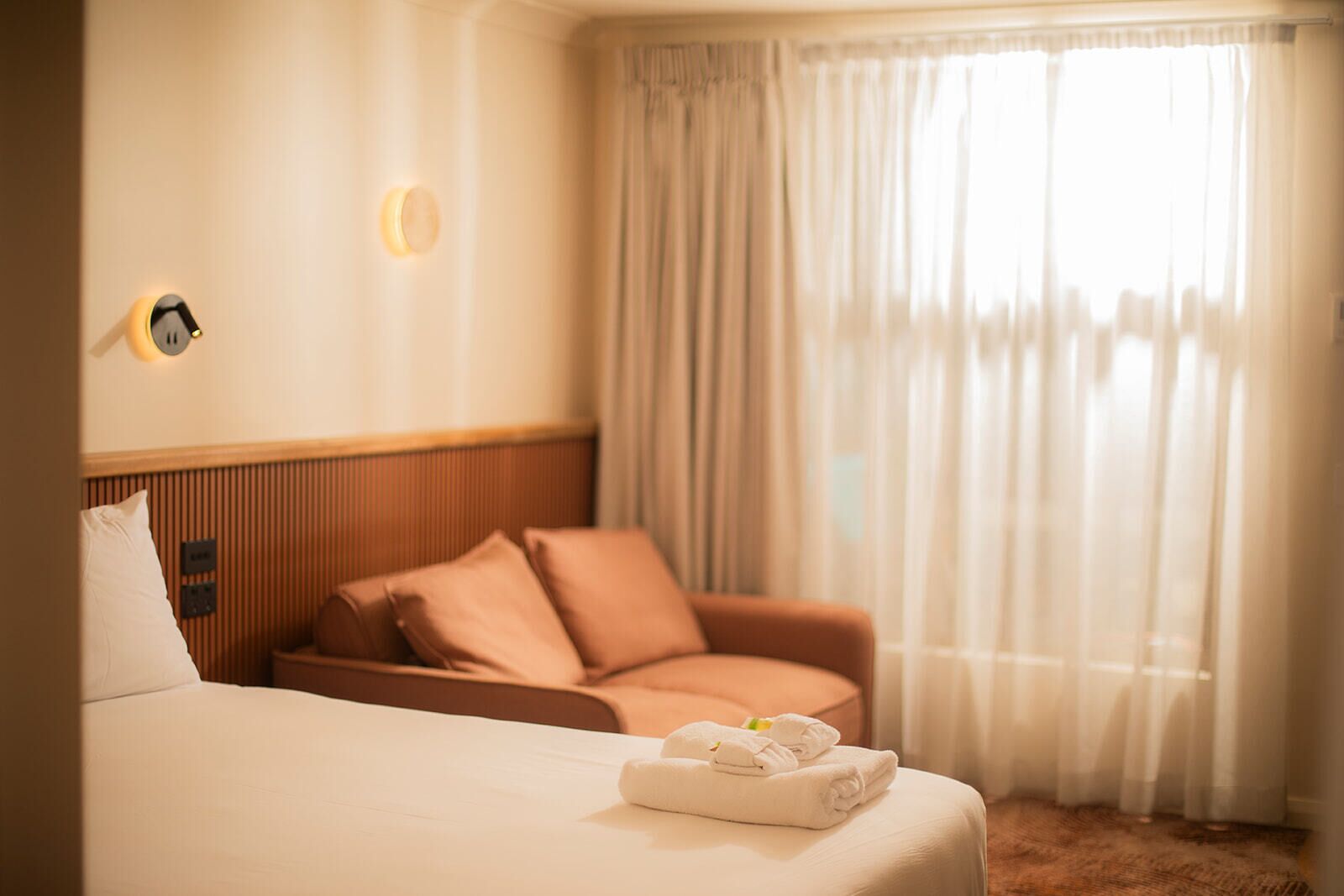 Una habitación de hotel con cama, cabecera de madera, sofá y ventana con cortinas.