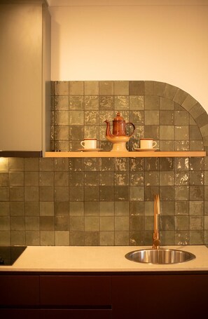 Una cocina con un lavamanos de acero inoxidable, un estante de madera con una tetera y tazas, y un salpicadero de azulejos.