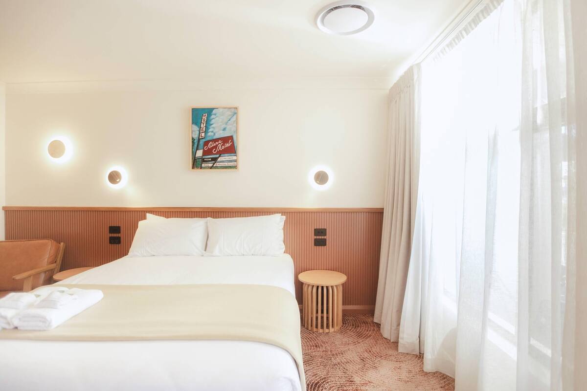 Una camera d'albergo con un letto, un tavolino rotondo in legno, una sedia, una lampada a parete e una finestra con tende leggere.