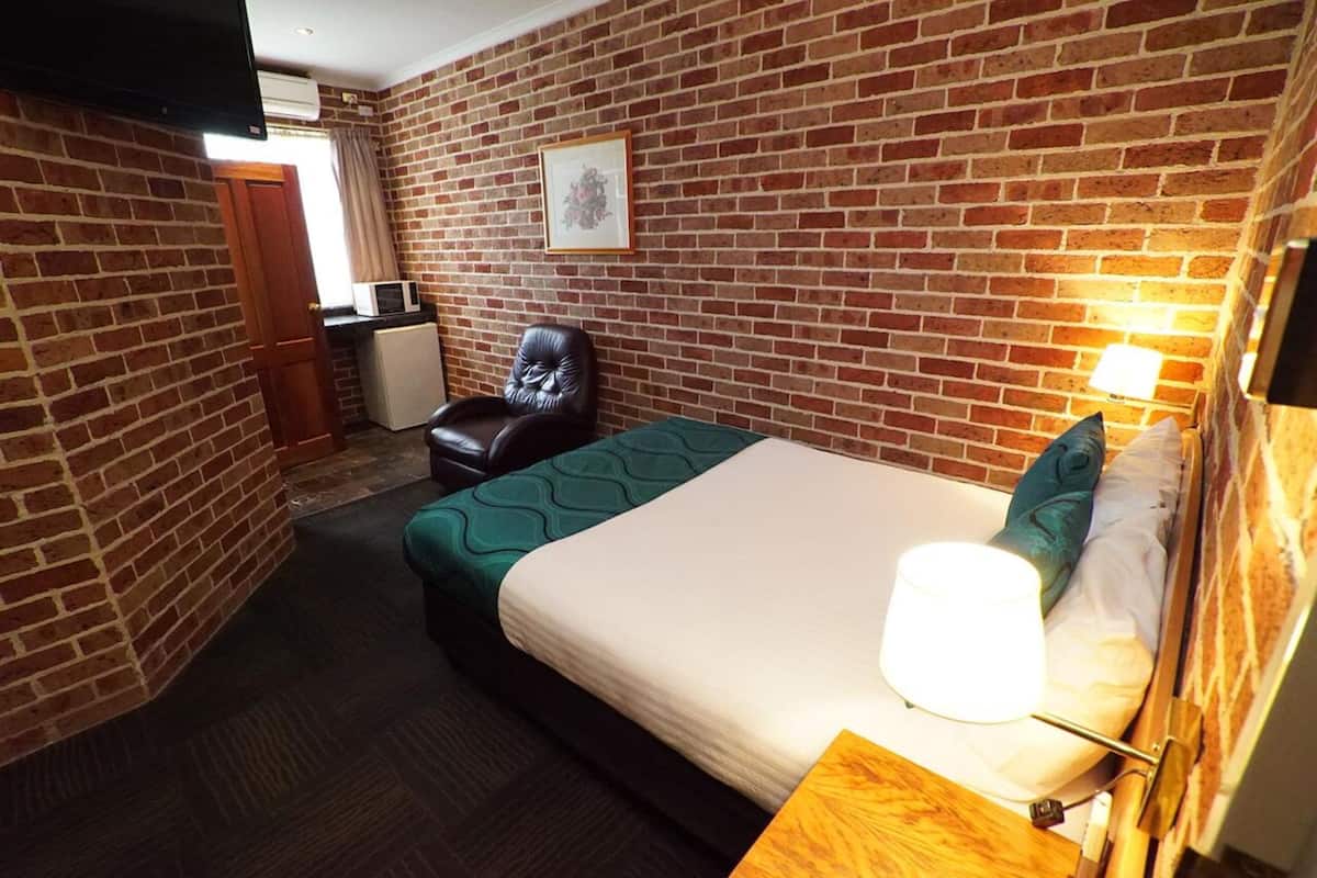 A hotel room with a brick wall, a bed with green bedding, a leather chair, a bedside table with a lamp, and a TV mounted on the wall.