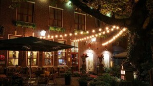 Un espacio al aire libre con mesas y sillas, un edificio de ladrillo con varias ventanas y luces colgantes.