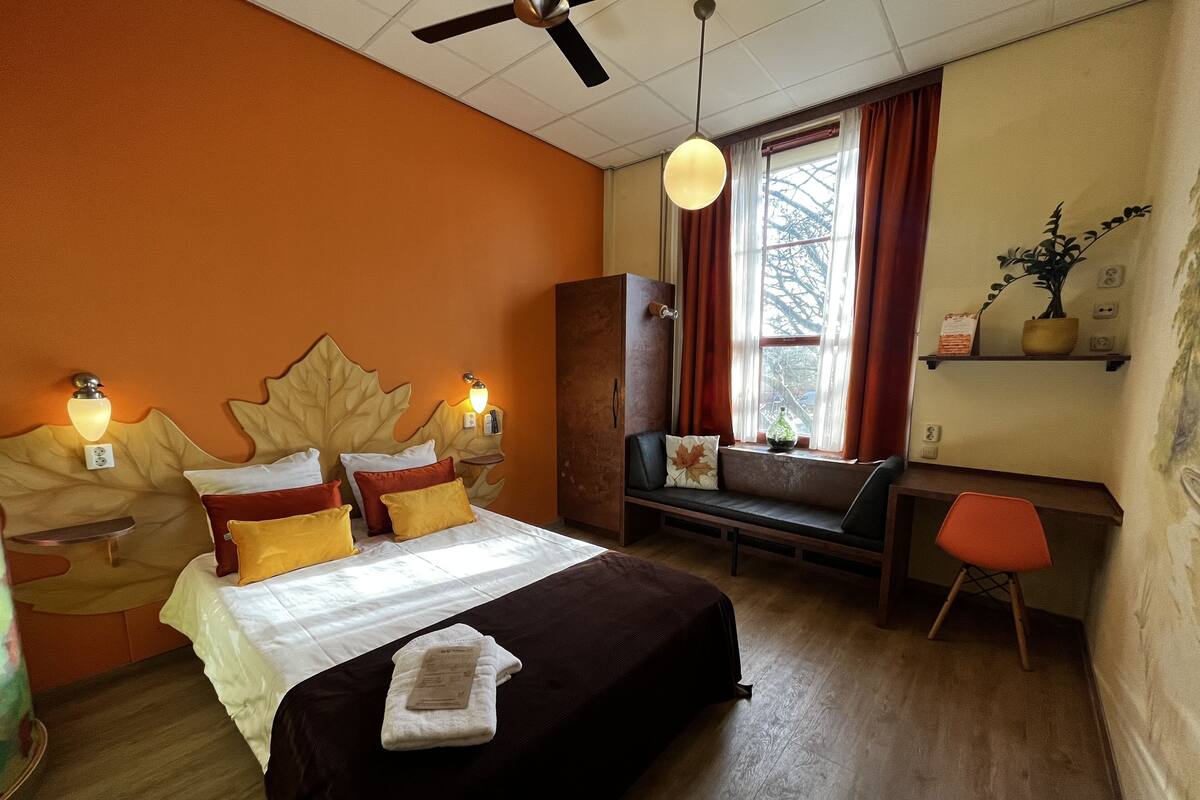 Ein Hotelzimmer mit einem großen Bett, einem Schreibtisch mit Stuhl, einem Fenster mit Vorhängen und einem Deckenventilator.