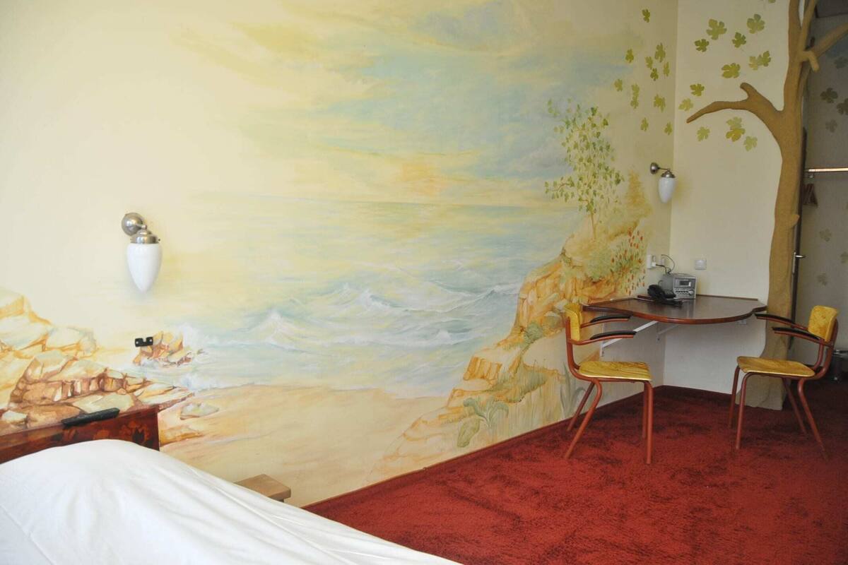 Ein Zimmer mit Wandgemälde einer Strandansicht, einem Bett, einem Schreibtisch mit zwei Stühlen und einem roten Teppich.