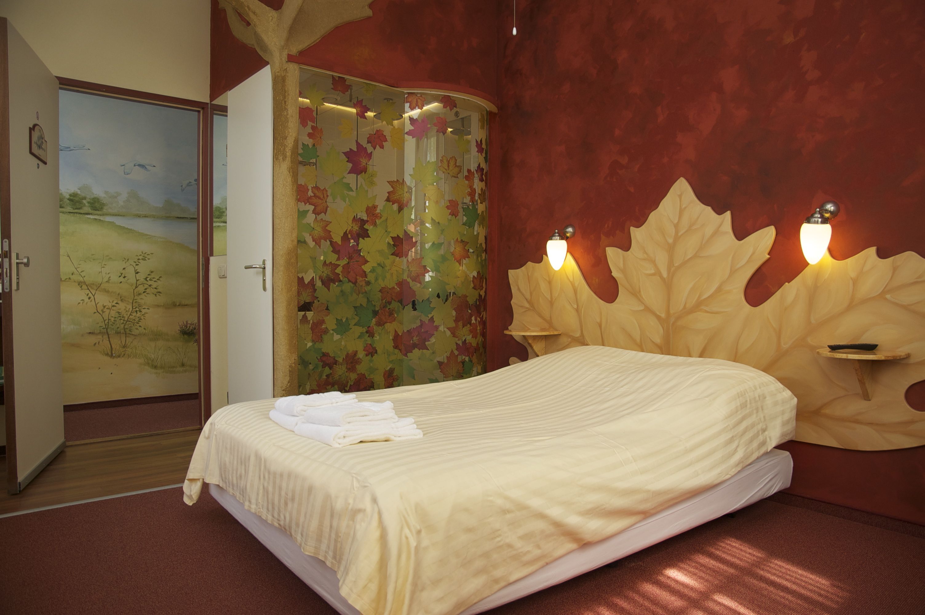 Una cama bien hecha con un edredón blanco y toallas dobladas, un cabecero en forma de hoja con luces empotradas en la pared, y una puerta con un mural de temática natural.
