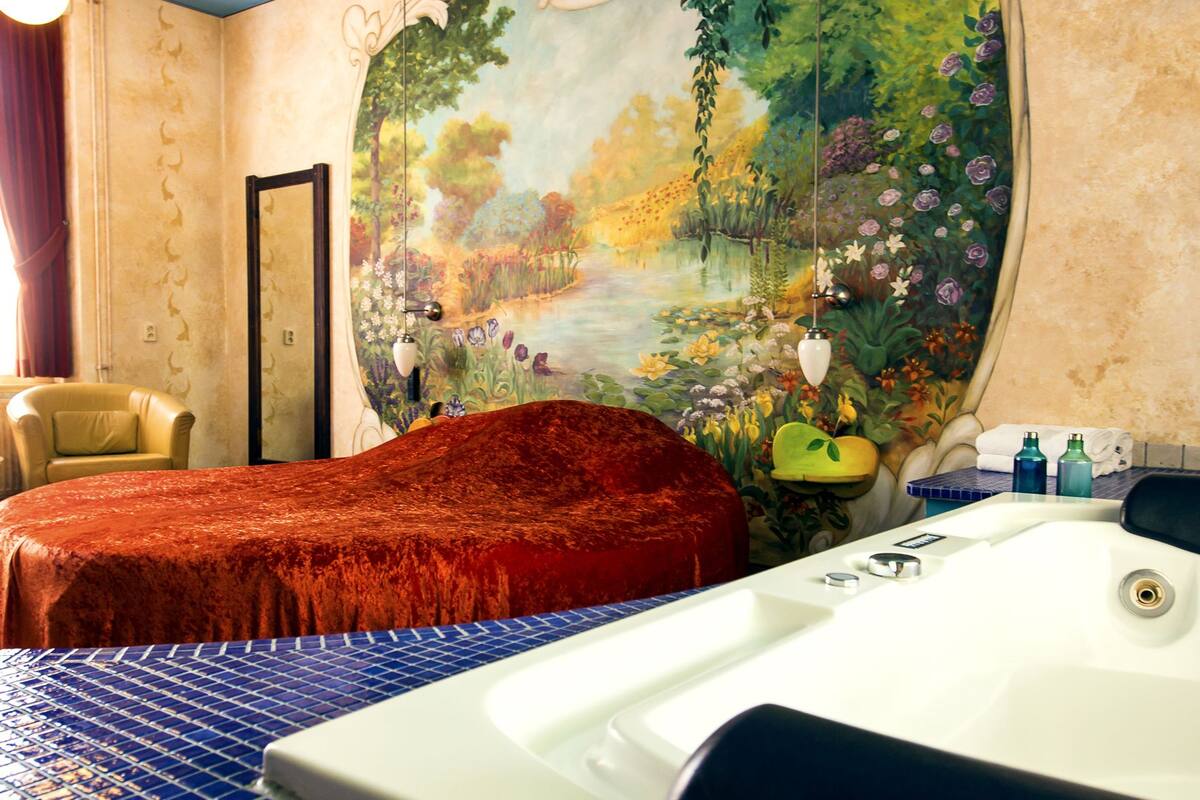 Ein Hotelzimmer mit einem großen Bett, einer Badewanne, einem Sessel und einem Wandgemälde mit einem Garten.