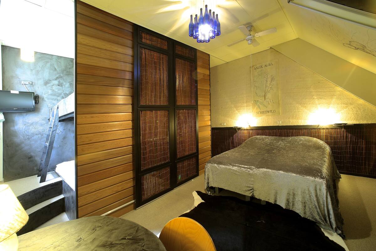 Ein Zimmer mit einem Bett, das mit einer Decke bezogen ist, einer Holzwand, einem Fenster mit Bambusvorhängen und einer Wandleuchte.