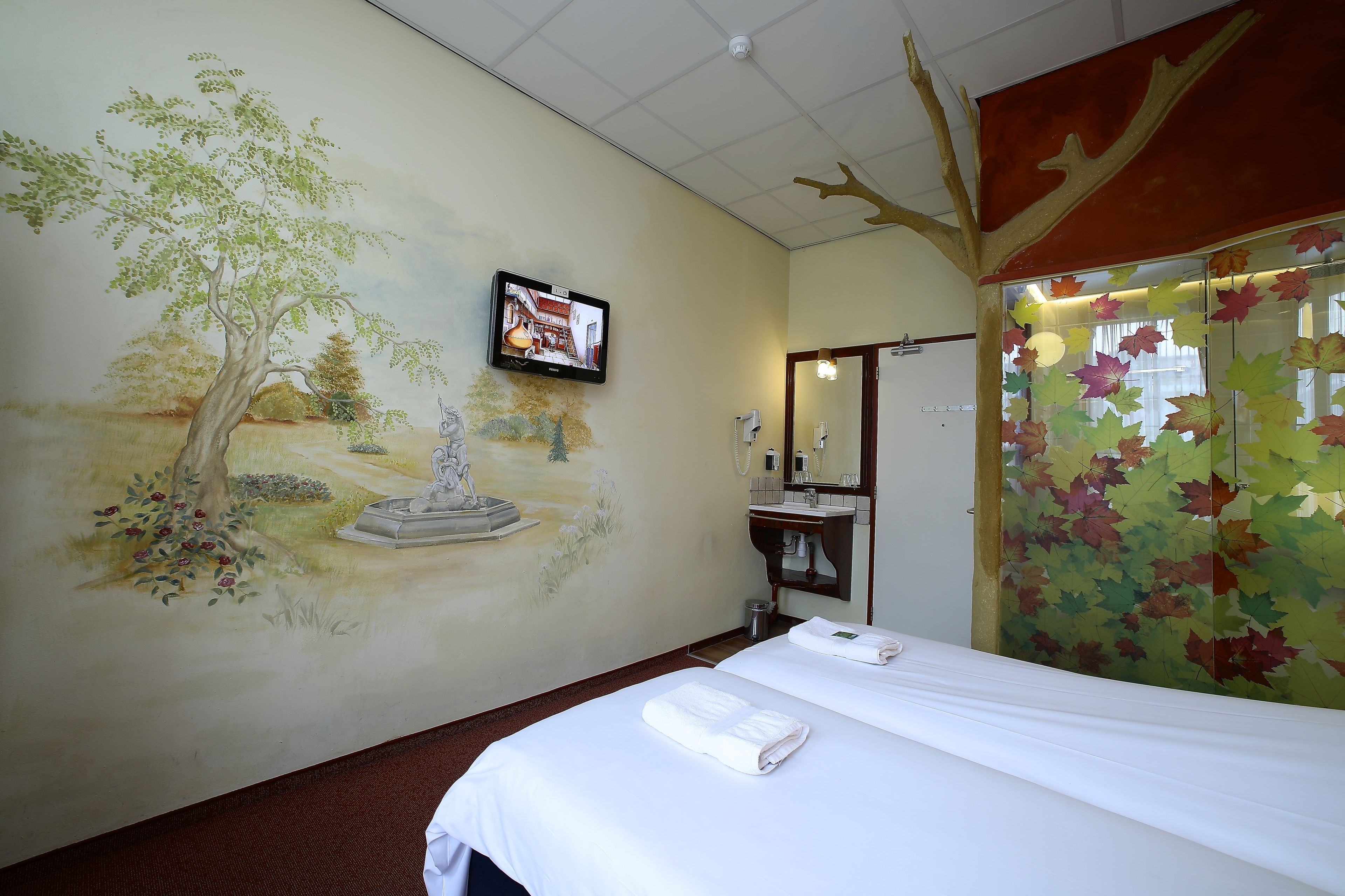 Une chambre d’hôtel avec une fresque représentant un arbre et une statue, un lit avec des serviettes, une télévision, un miroir et une salle de bain avec un lavabo et un miroir.