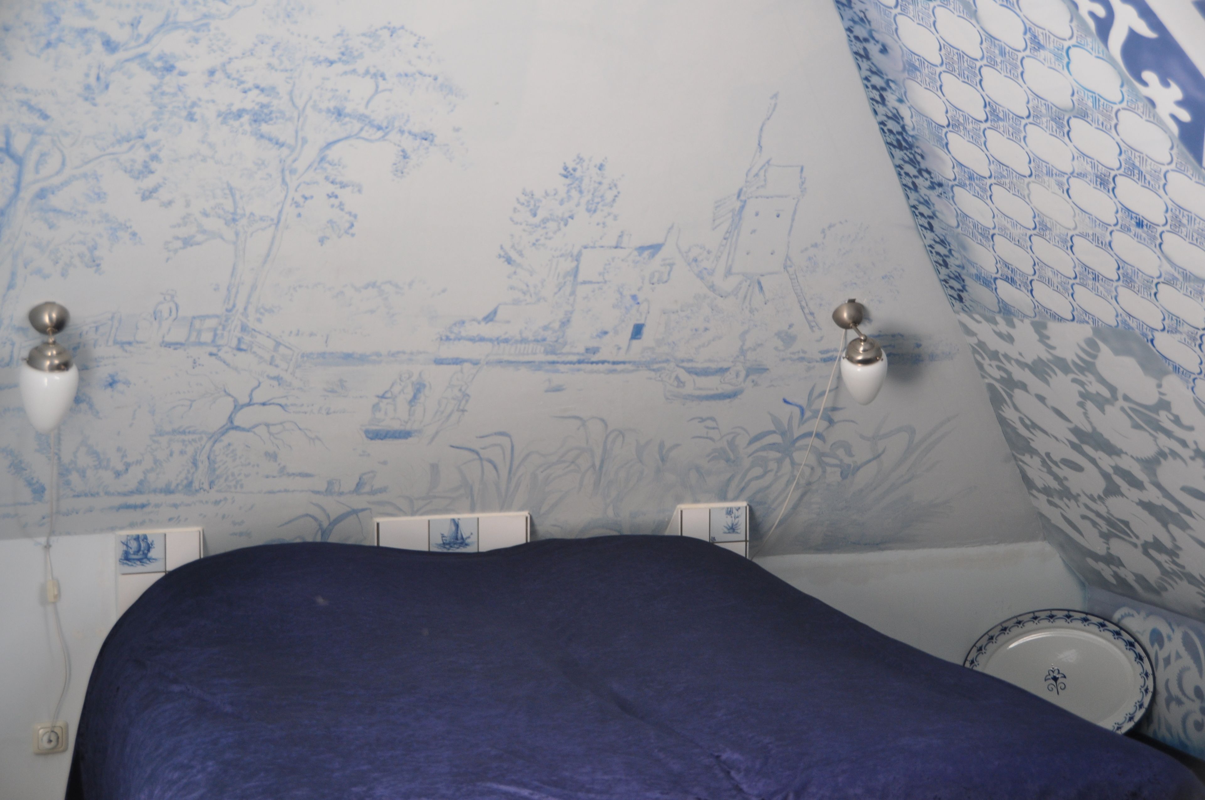 Une chambre à coucher avec un lit, un mur orné d’un motif bleu et blanc, et une petite table sur laquelle se trouve une assiette.