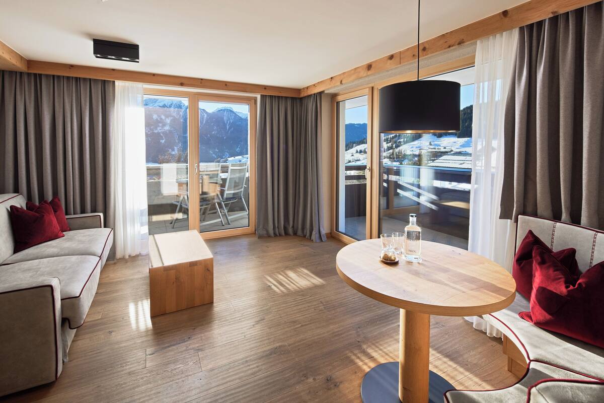 A modern hotel room with a wooden floor, a round table, a sofa, and a balcony with a view of mountains.