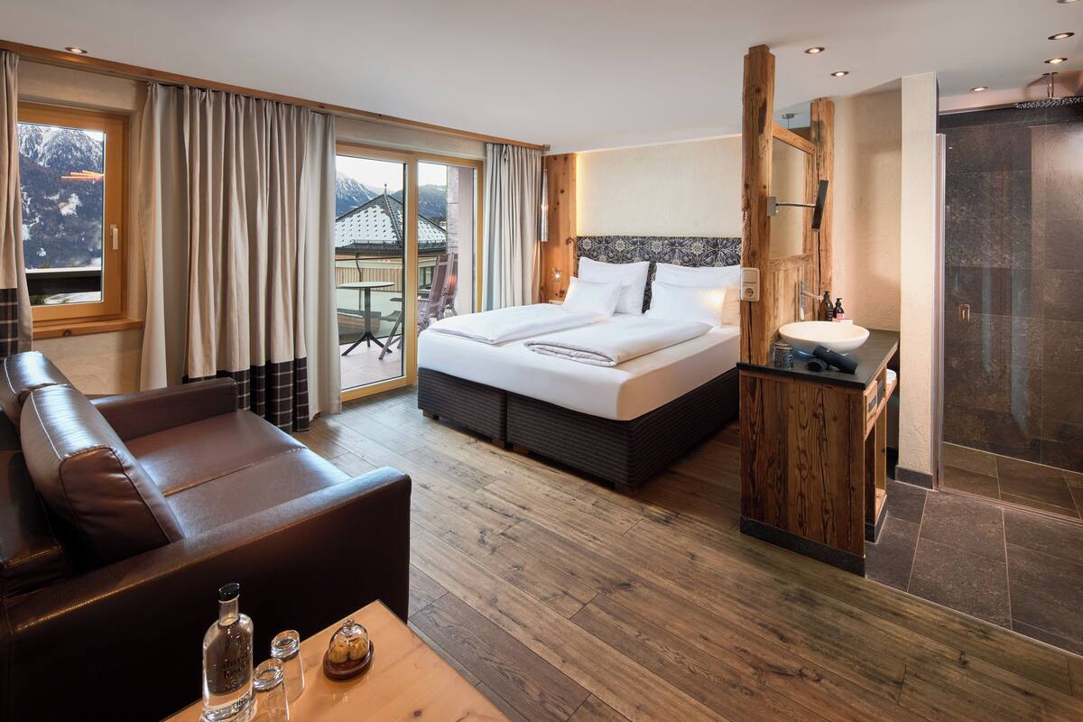 A hotel room with a bed, a sofa, a wooden table, a bathroom with a shower, and a view of mountains.