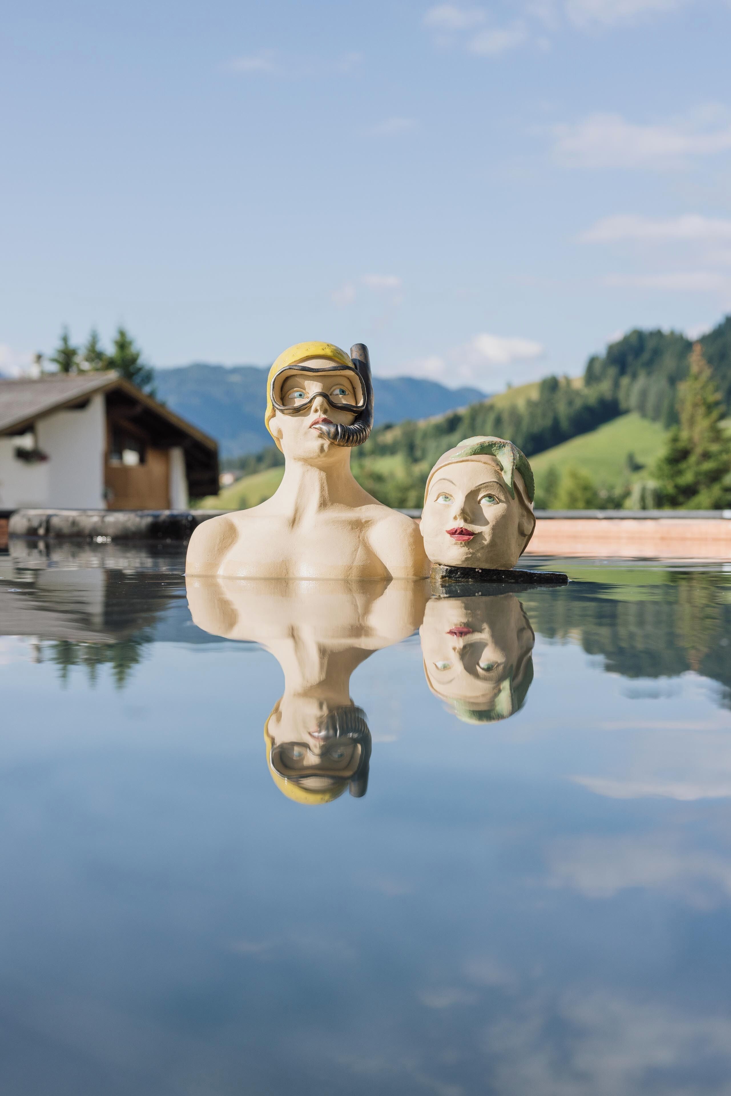 A reflective pool with two sculptures of a man and a woman, a wooden cabin, and a mountainous landscape in the background.