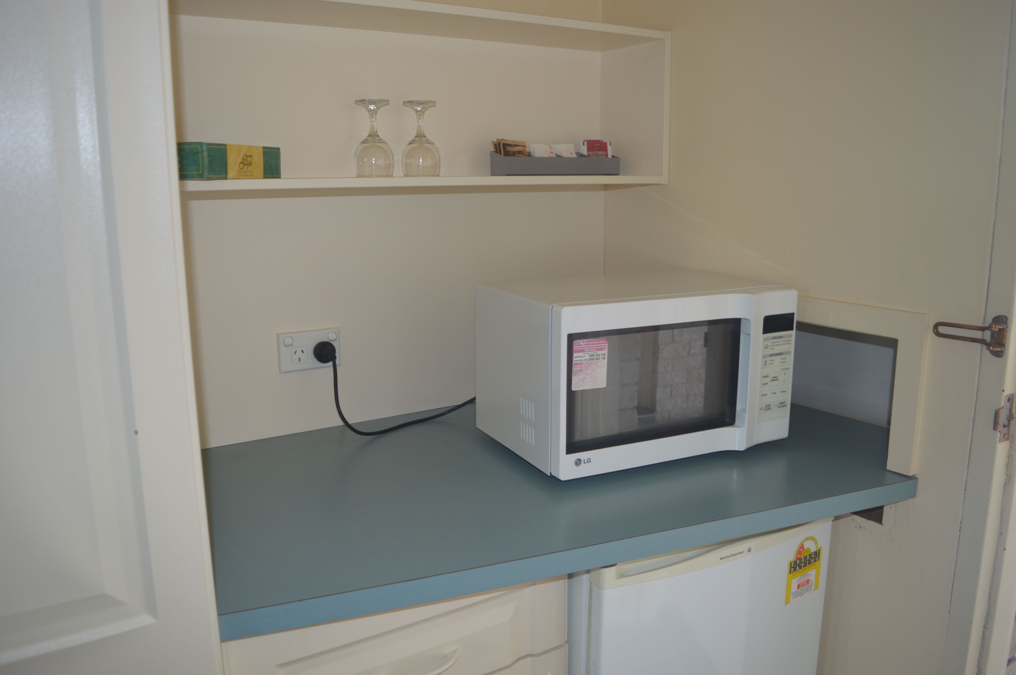 A microwave on a countertop with a shelf above it containing a few items.