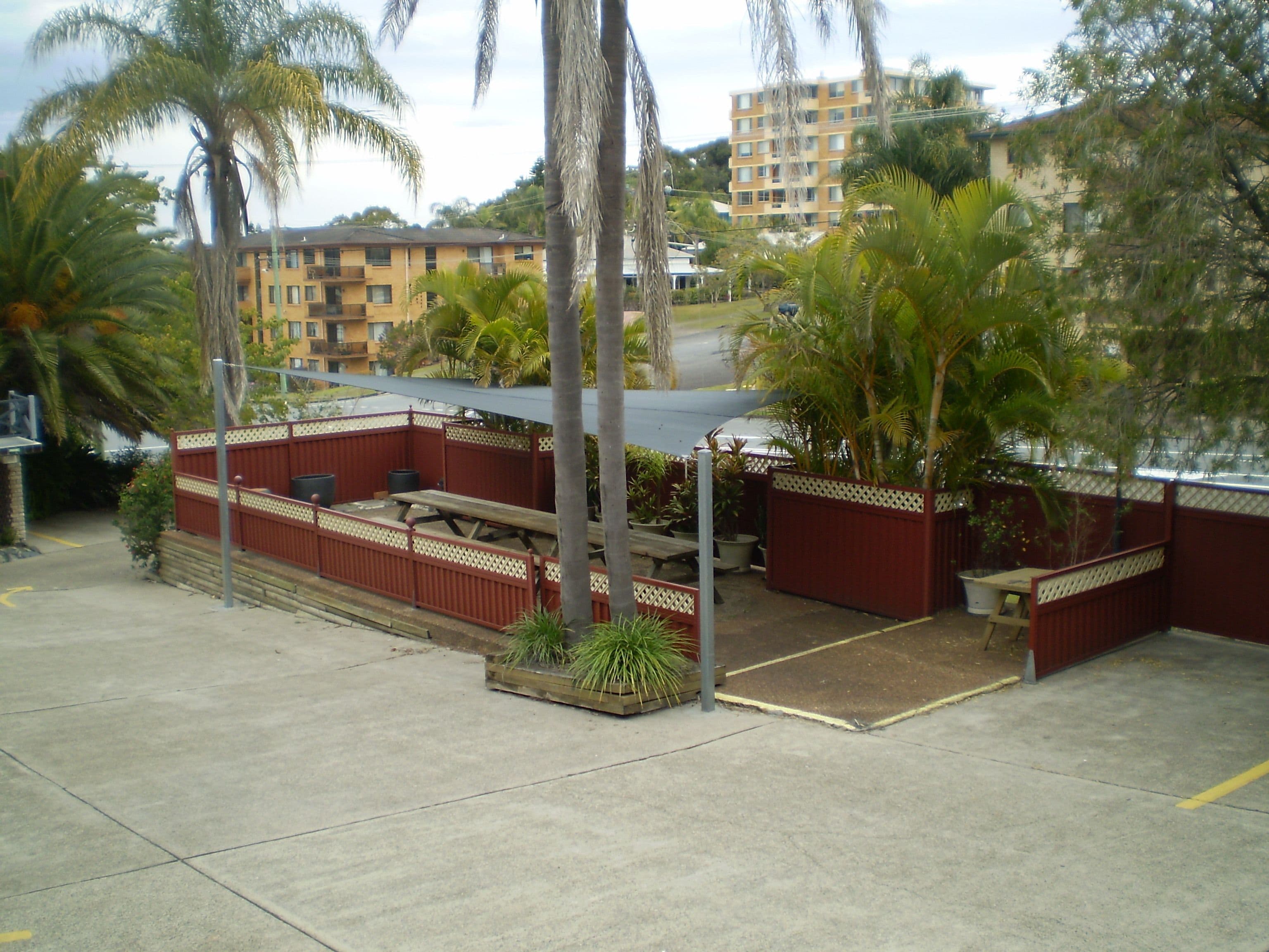 A covered outdoor seating area with benches and tables, surrounded by palm trees and a red fence.