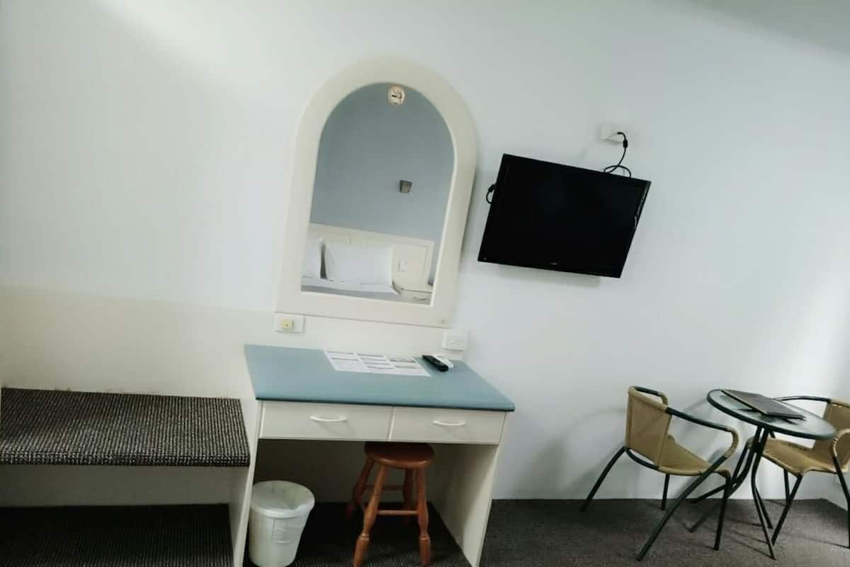 A compact hotel room with a desk, a chair, a small stool, a television mounted on the wall, and a trash bin.