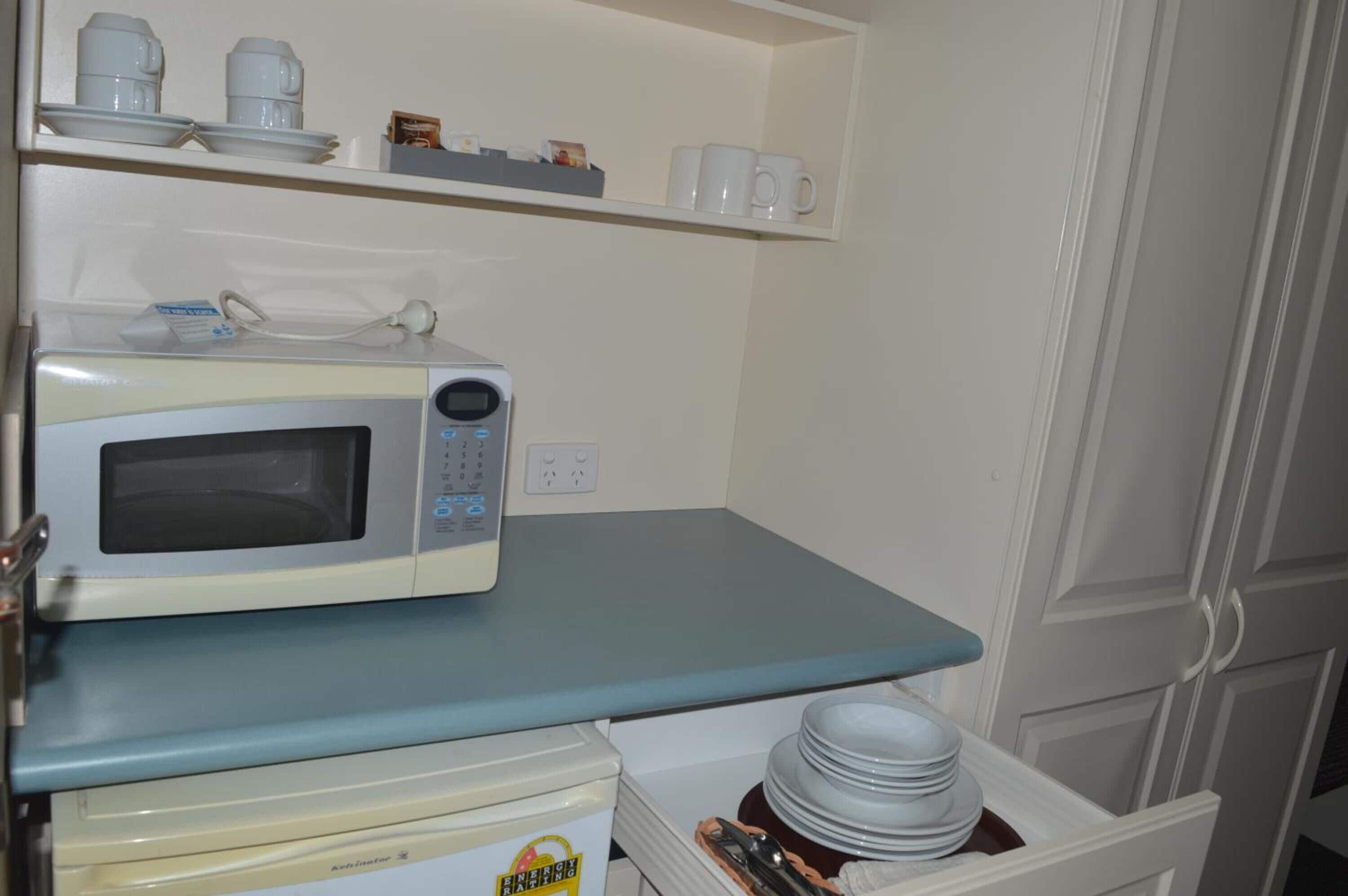 Une cuisine avec un four à micro-ondes posé sur un plan de travail bleu, une étagère avec des tasses et des assiettes, et un tiroir contenant des assiettes empilées.
