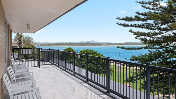 A balcony with white chairs, a black railing, and a view of the sea and distant mountains. - El Motor Inn (Port Macquarie)