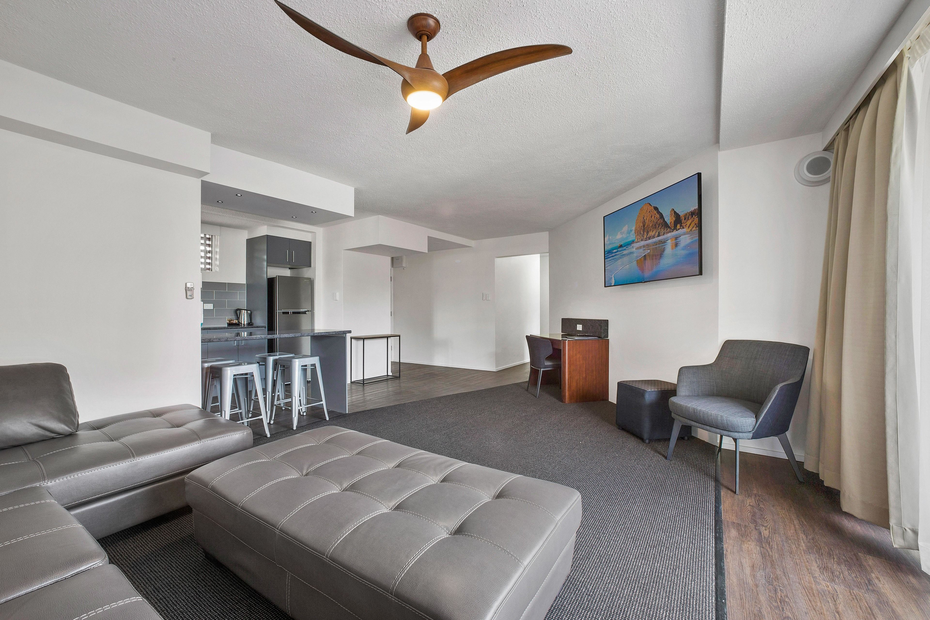 A modern living room with a grey sectional sofa, a ceiling fan, a dining area with a kitchen in the background, and a painting on the wall.