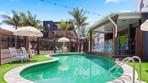 A swimming pool with a curved edge, surrounded by a stone deck and a grassy area. There are lounge chairs and umbrellas, and a building with balconies in the background. - El Motor Inn (Port Macquarie)