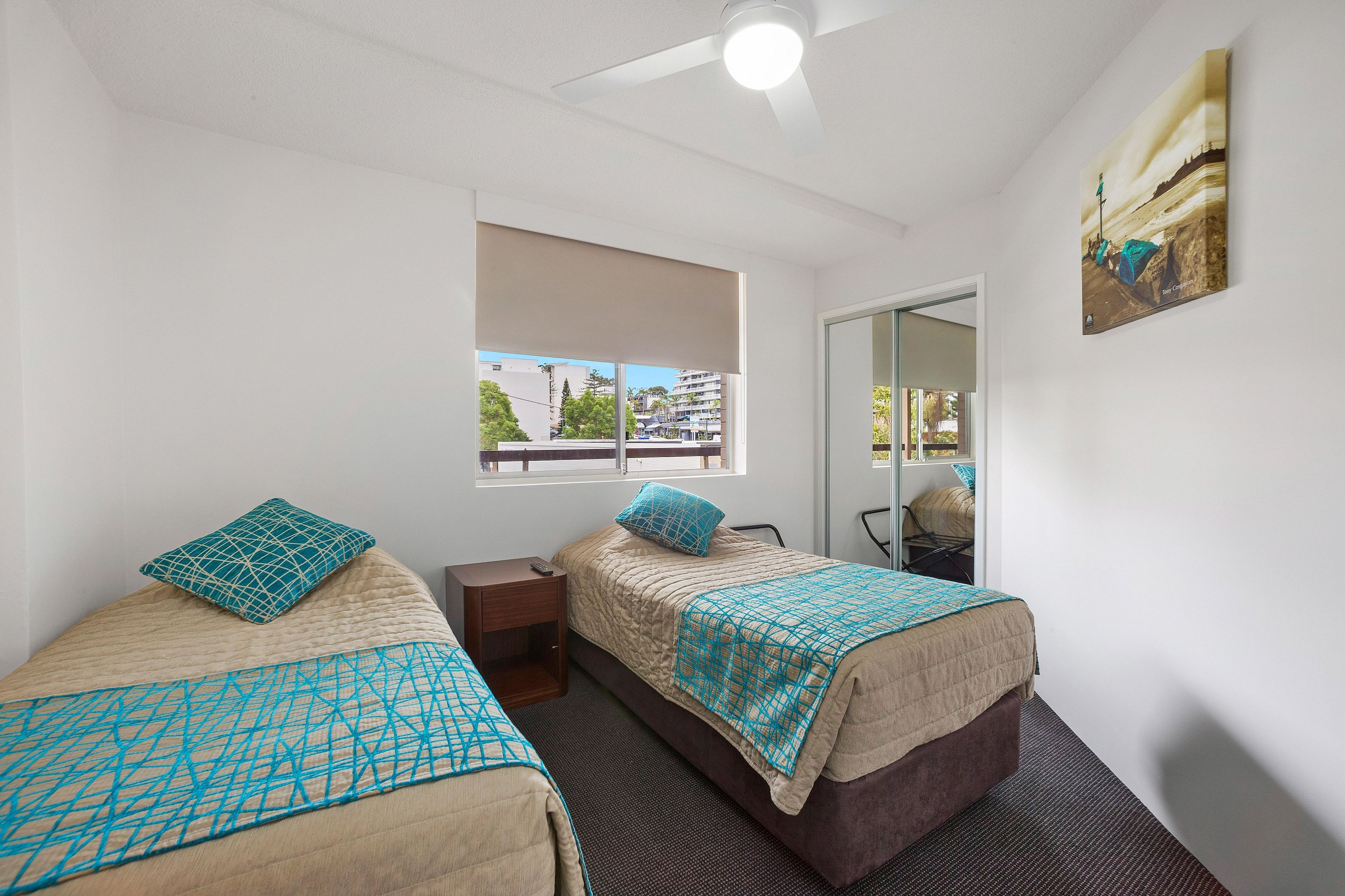 A hotel room with two beds, a window with a view, a ceiling fan, and a painting on the wall.