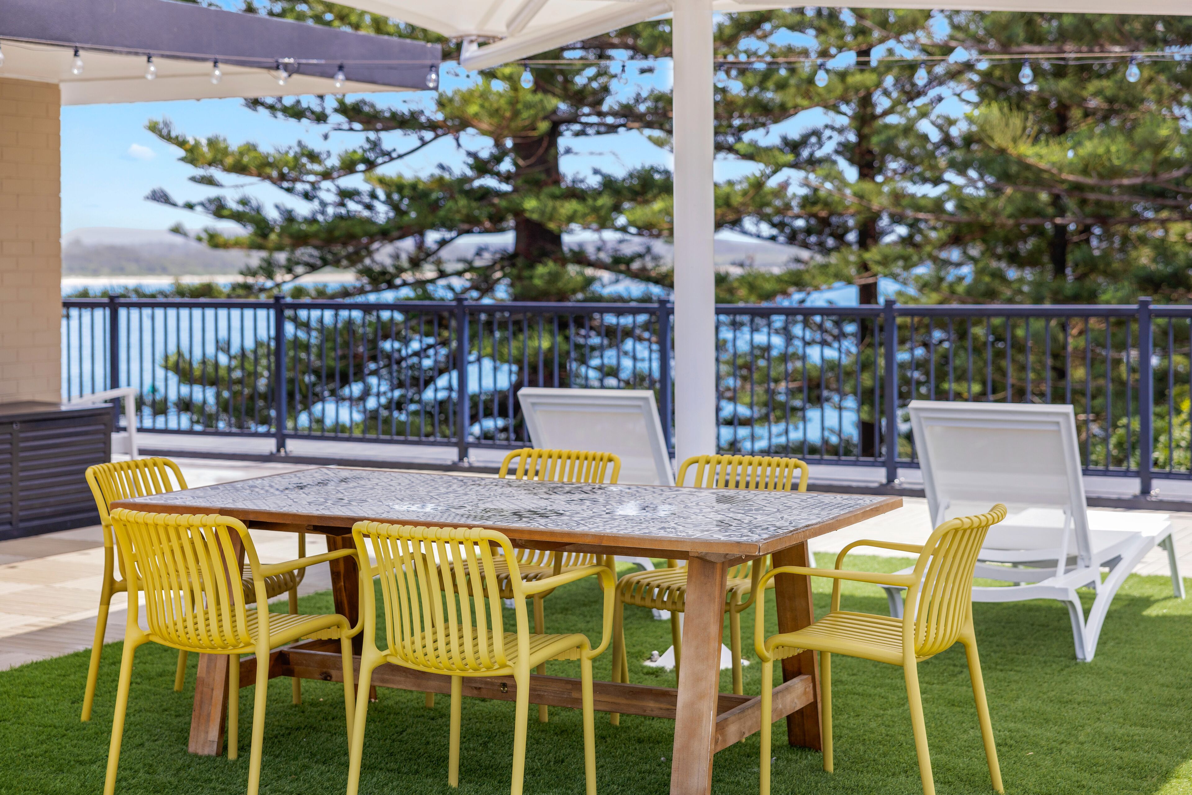 An outdoor patio with a wooden table and yellow chairs, overlooking a body of water and trees.