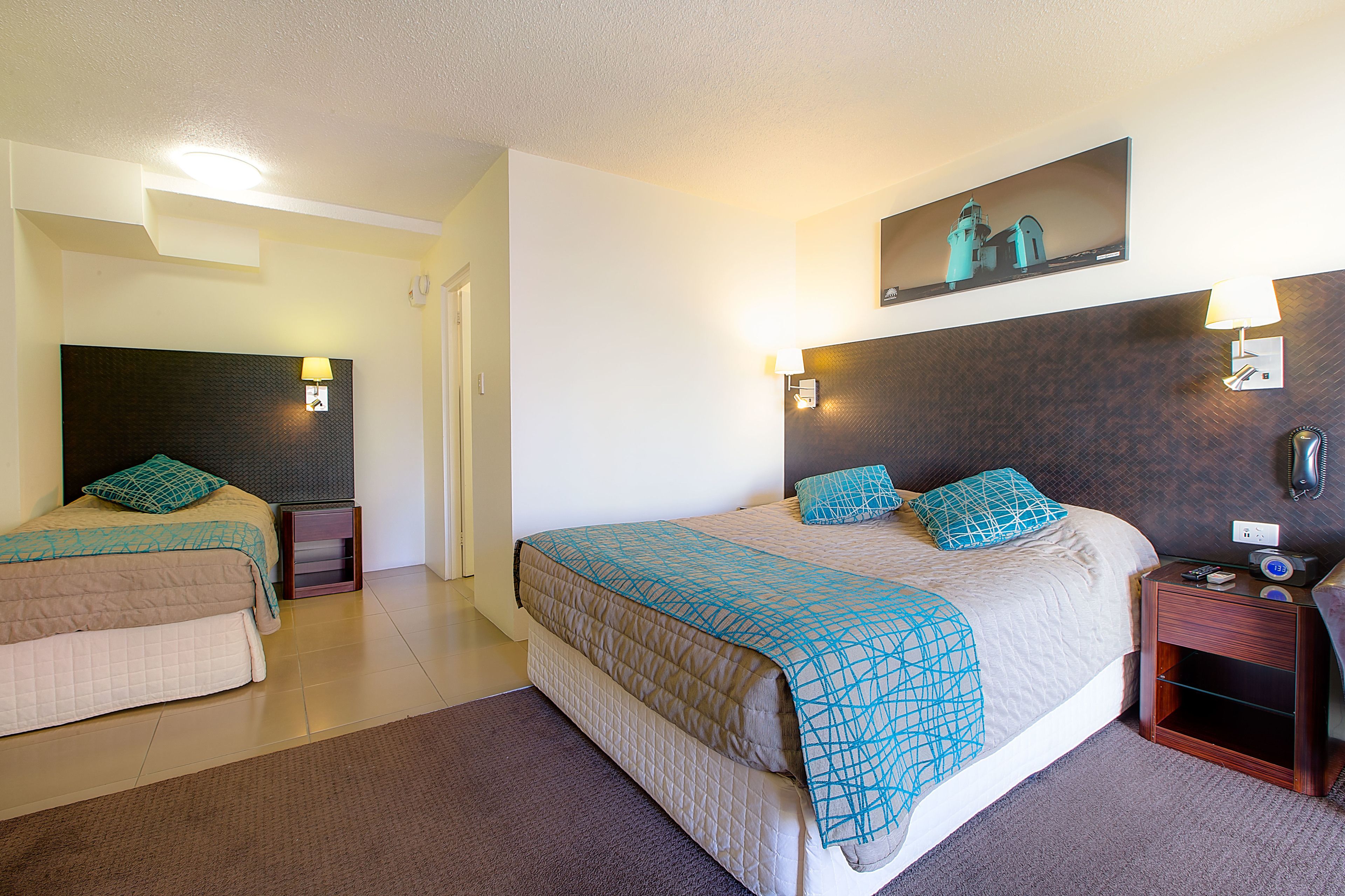 Habitación de hotel con dos camas, una cama grande con colcha estampada, una cama más pequeña con colcha lisa, una mesita de noche con lámpara, un teléfono montado en la pared y un cuadro enmarcado sobre la cama grande.