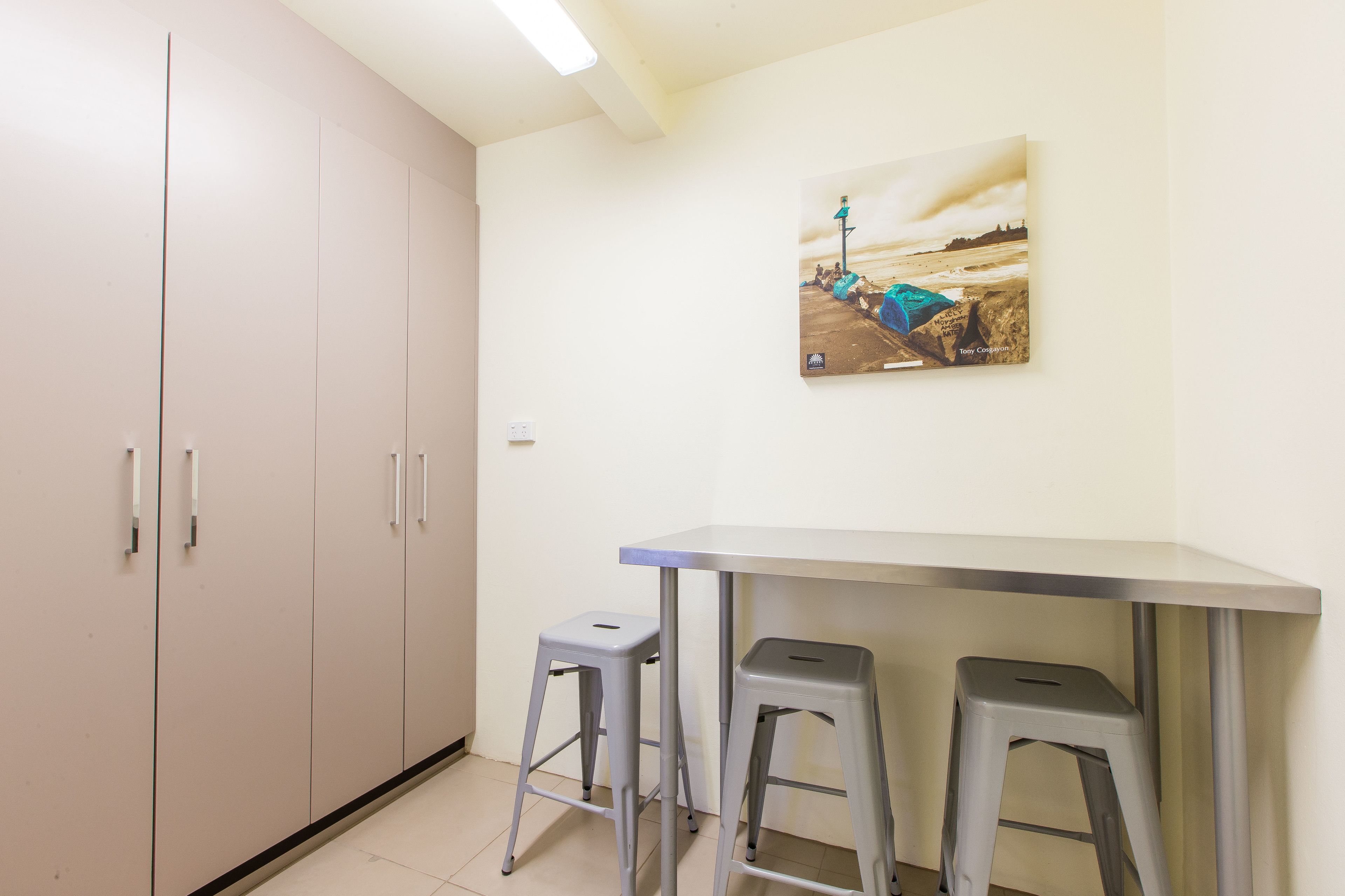 Una habitación con pared blanca, un cuadro de una playa, una mesa blanca y dos taburetes grises.