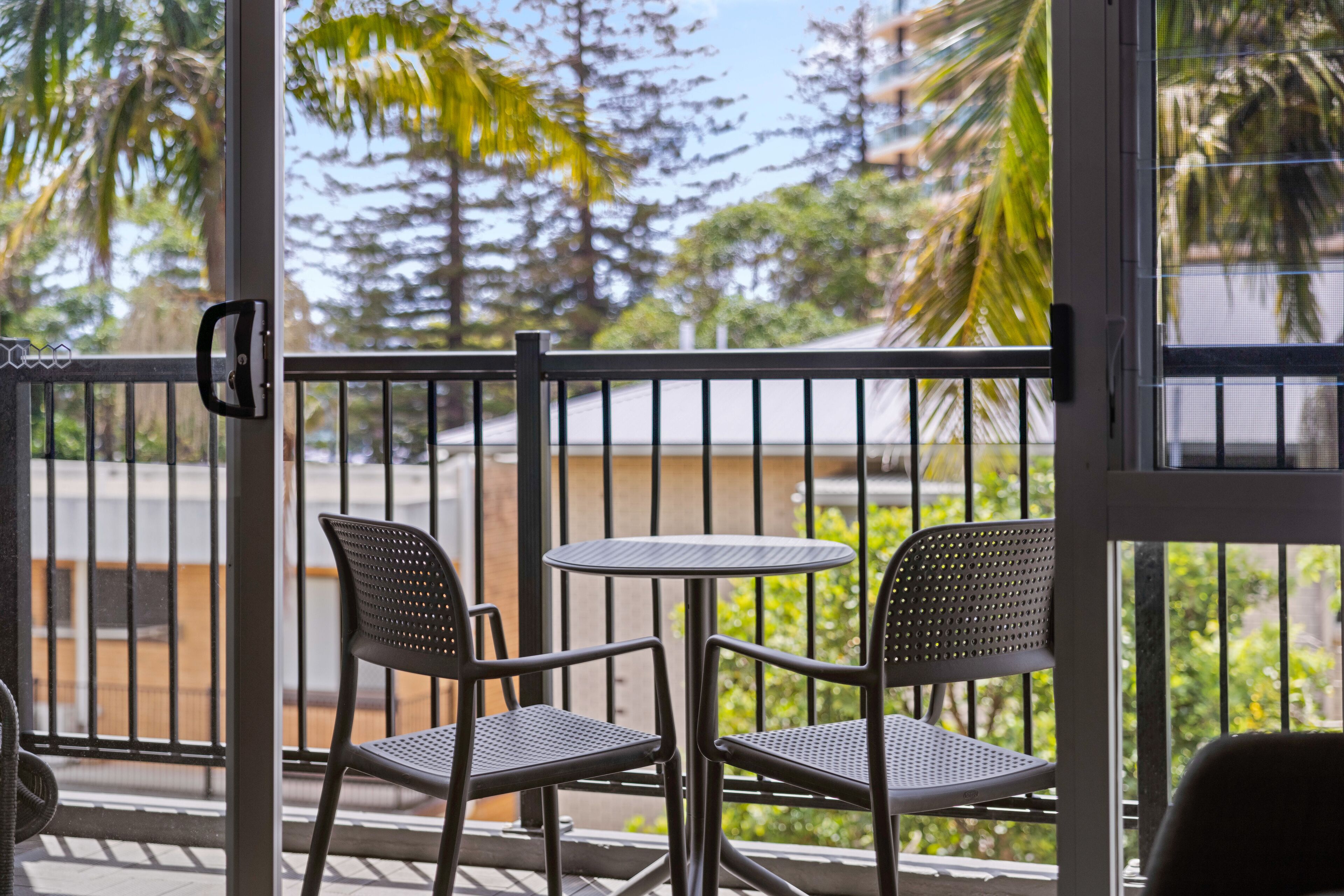 A balcony with two chairs and a small table, overlooking a street with palm trees and a building in the background.