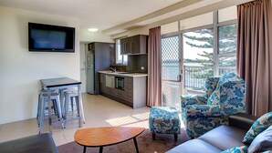 A modern living room with a sofa, ottoman, and a small wooden table. A kitchen area is visible with cabinets and a microwave. A flat-screen TV is mounted on the wall. - El Motor Inn (Port Macquarie)