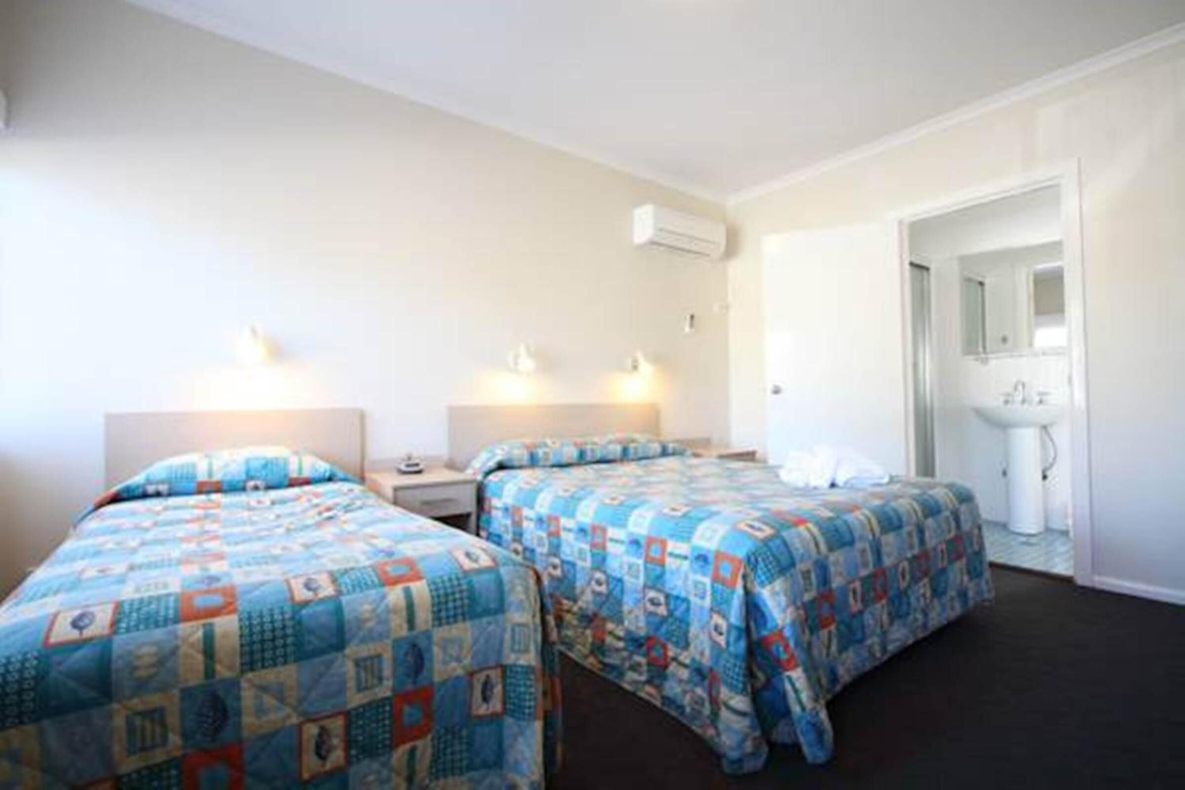 A hotel room with two beds, a bedside table, a nightstand, a mirror, and a bathroom with a sink and mirror.