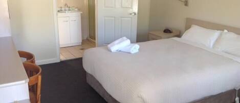 Habitación de hotel con cama, escritorio, silla, mesita de noche y baño visible a través de una puerta abierta.