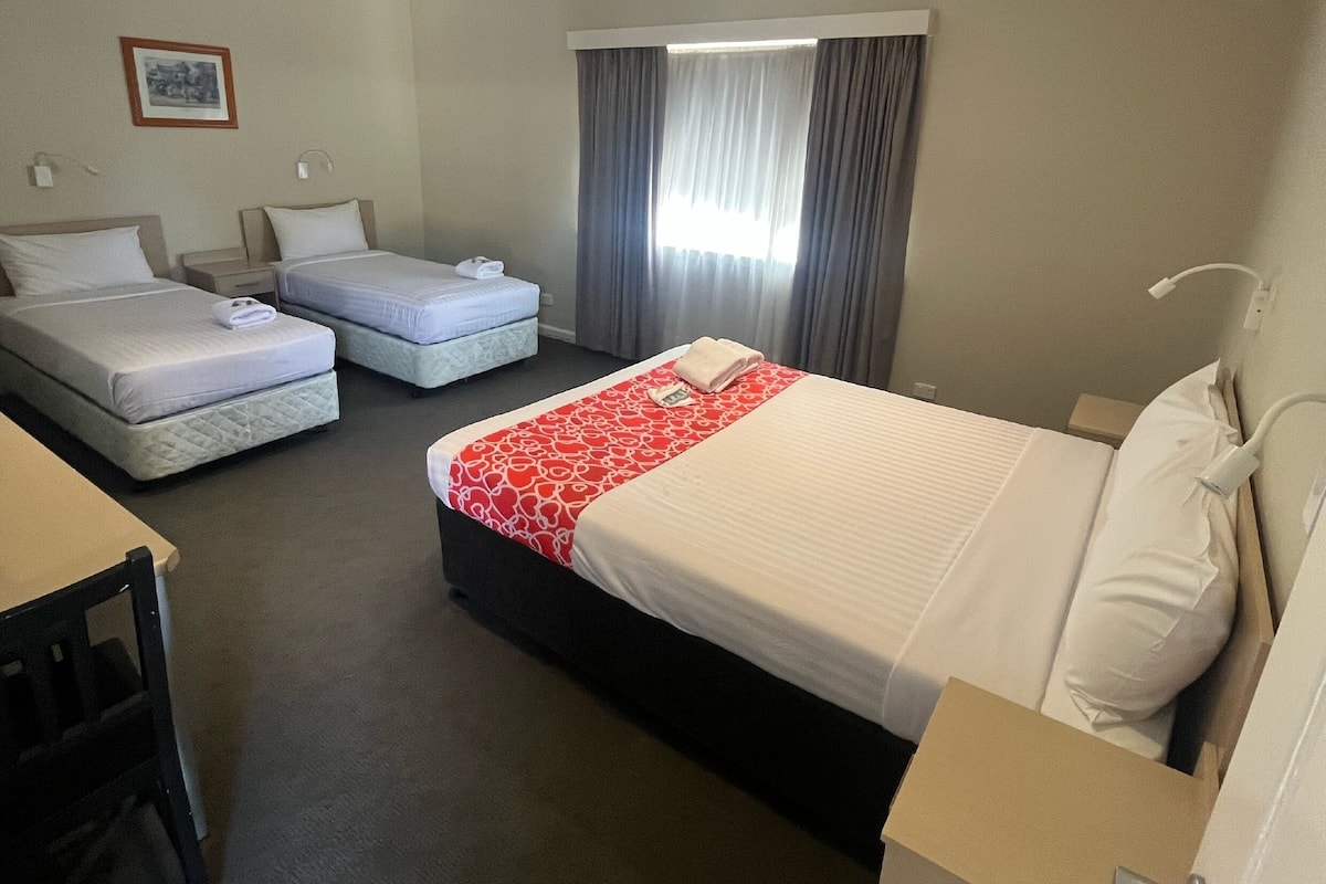 A hotel room with two single beds, a larger bed, a desk, and a chair.