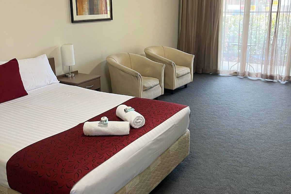 A hotel room with a large bed, two chairs, a nightstand, a lamp, a framed picture on the wall, and a window with curtains.