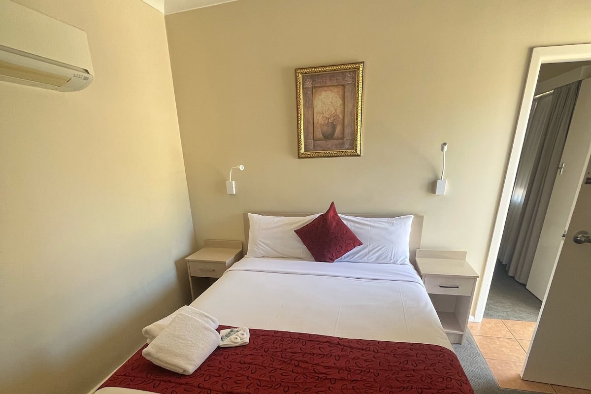A hotel room with a bed, a red throw blanket, a white pillow, and a framed picture on the wall.