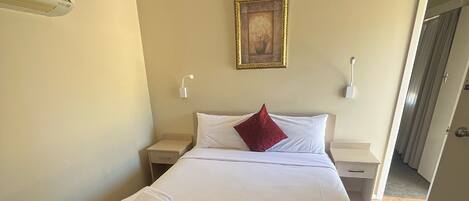 Habitación de hotel con una cama, una manta roja, un almohadón blanco y un cuadro en la pared.