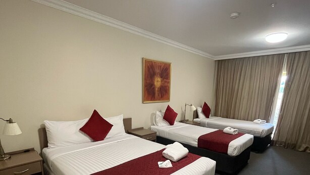 A hotel room with two beds, each with white linens and red pillows, a red throw blanket, a painting on the wall, and a large window with curtains. - Gabriel Motor Inn (Gundagai)