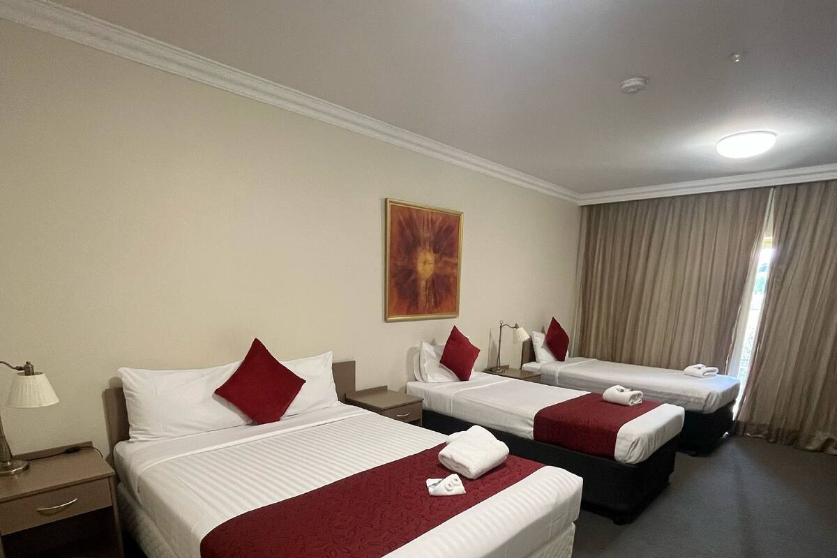 A hotel room with two beds, each with white linens and red pillows, a red throw blanket, a painting on the wall, and a large window with curtains.