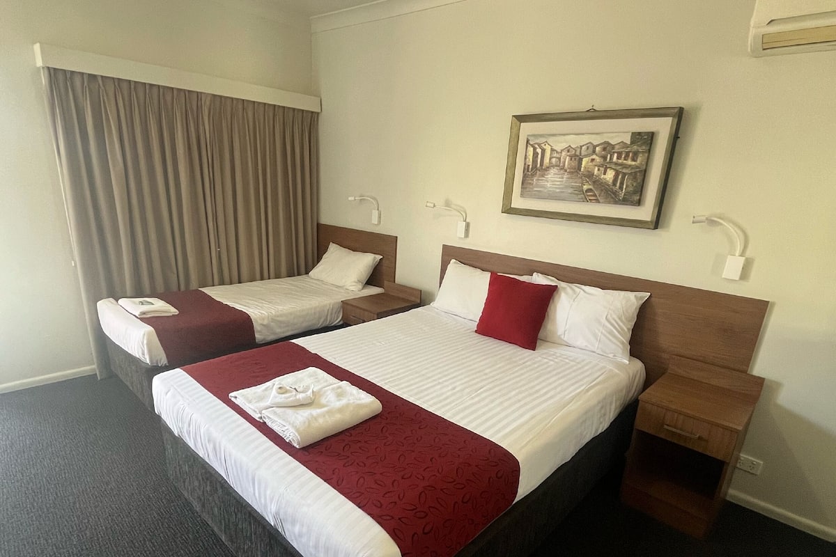 A hotel room with two single beds, a large bed, a wooden nightstand, a framed picture on the wall, and a window with curtains.
