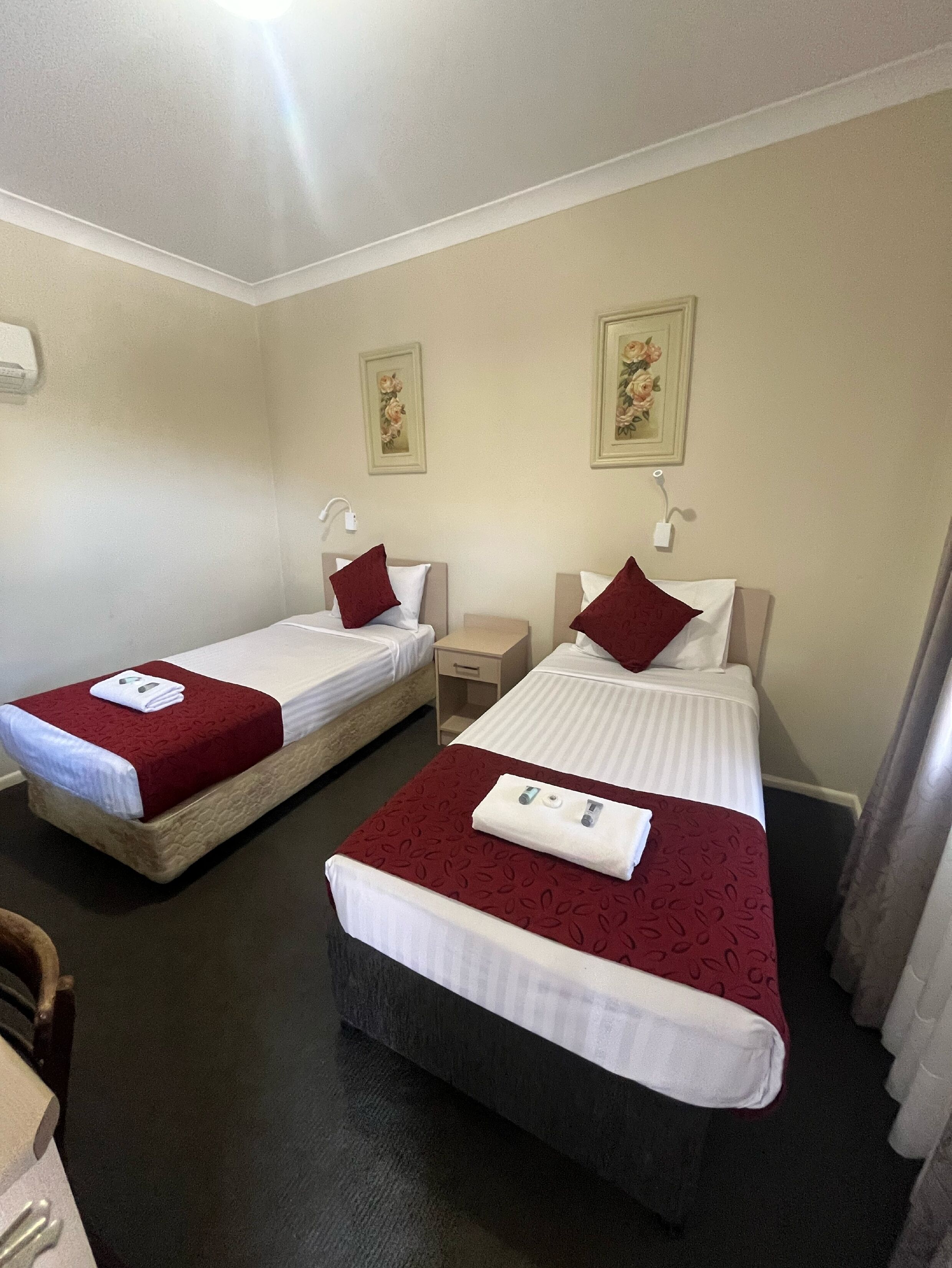Habitación de hotel con dos camas individuales, cada una con colcha roja y ropa de cama blanca, una mesita de noche con lámpara y una pequeña cómoda.