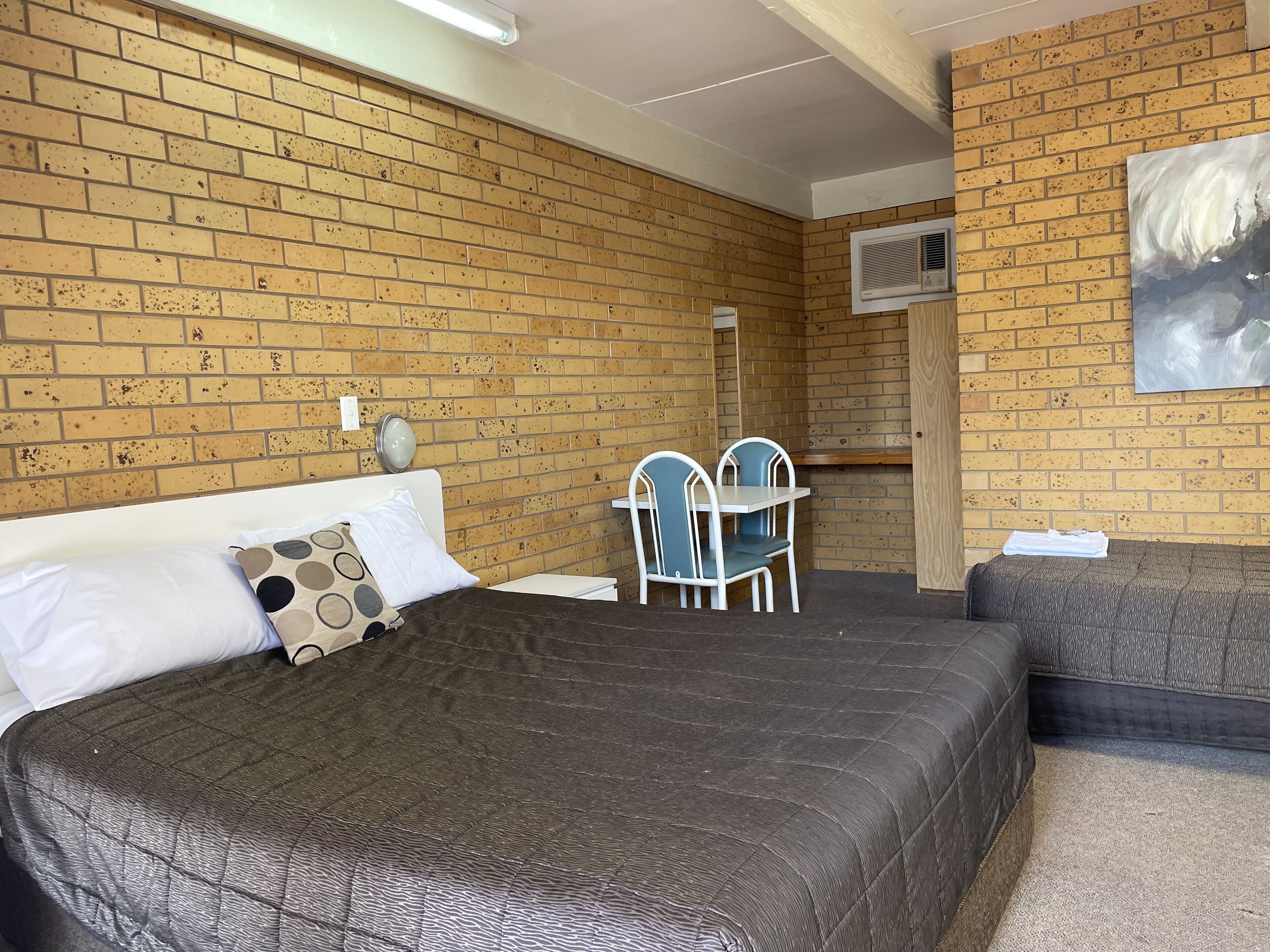 A room with a brick wall, a bed with a dark quilt, a small table with chairs, and a sofa.