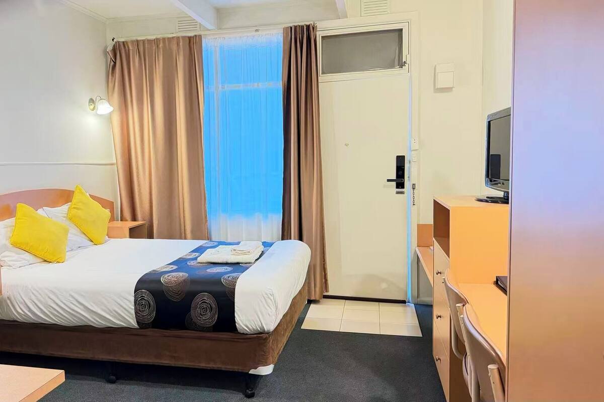 A hotel room with a bed, two bedside tables, a TV, and a window with curtains.