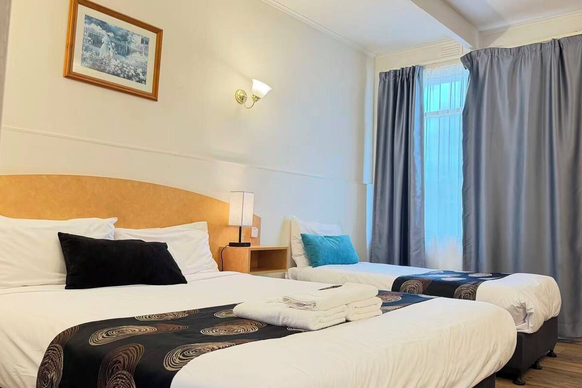 A hotel room with two beds, a nightstand, a lamp, a framed picture on the wall, and a window with curtains.