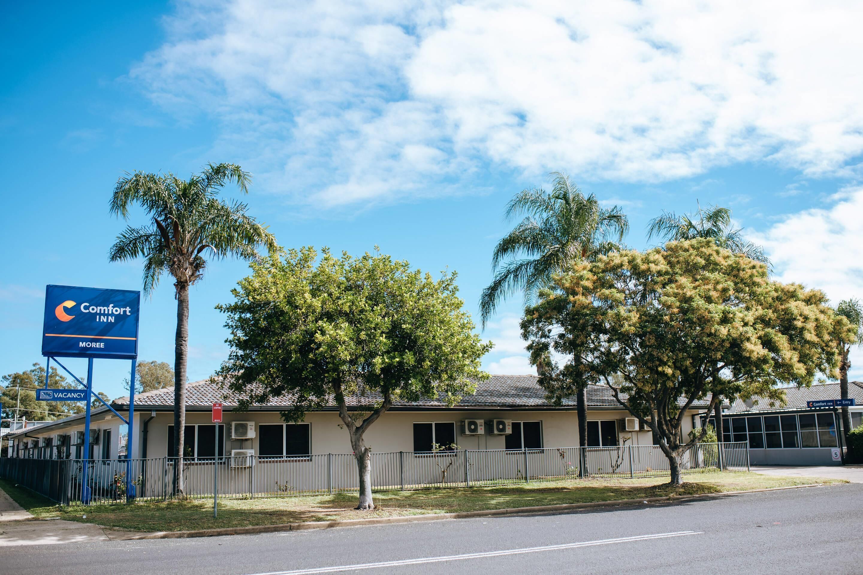 A Comfort Inn hotel with a fenced parking area, palm trees, and a clear blue sky.