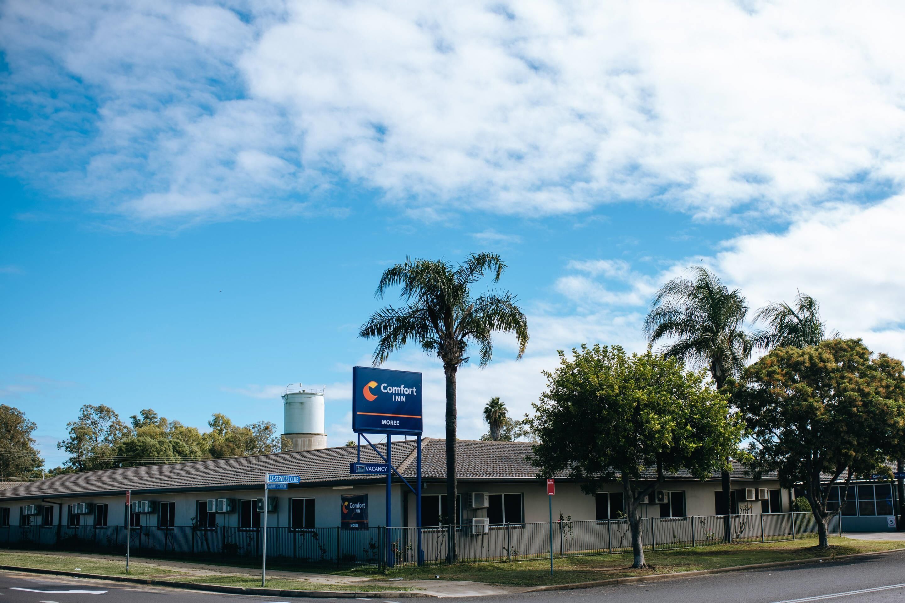 A motel with a signboard, fenced parking, and a water tower in the background.
