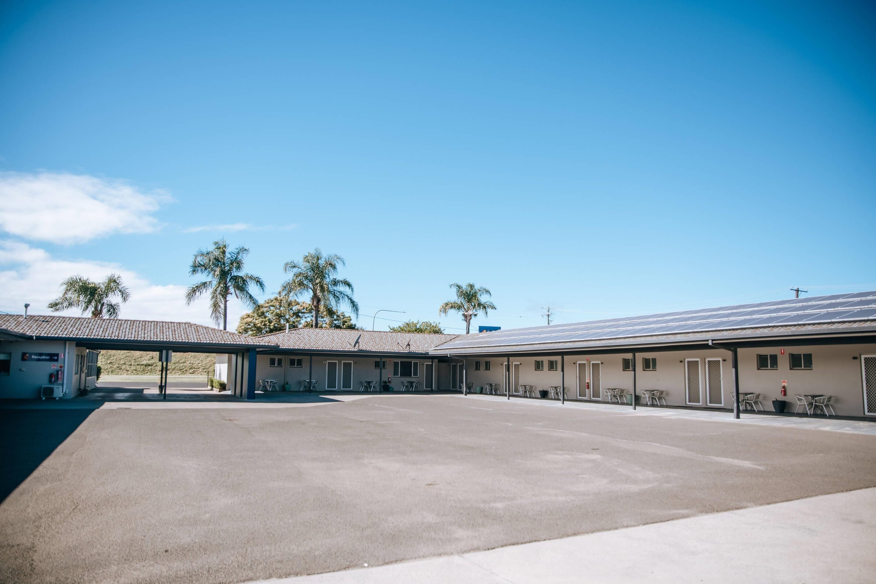 Un motel con área de estacionamiento techada, varias habitaciones y cielo azul despejado.