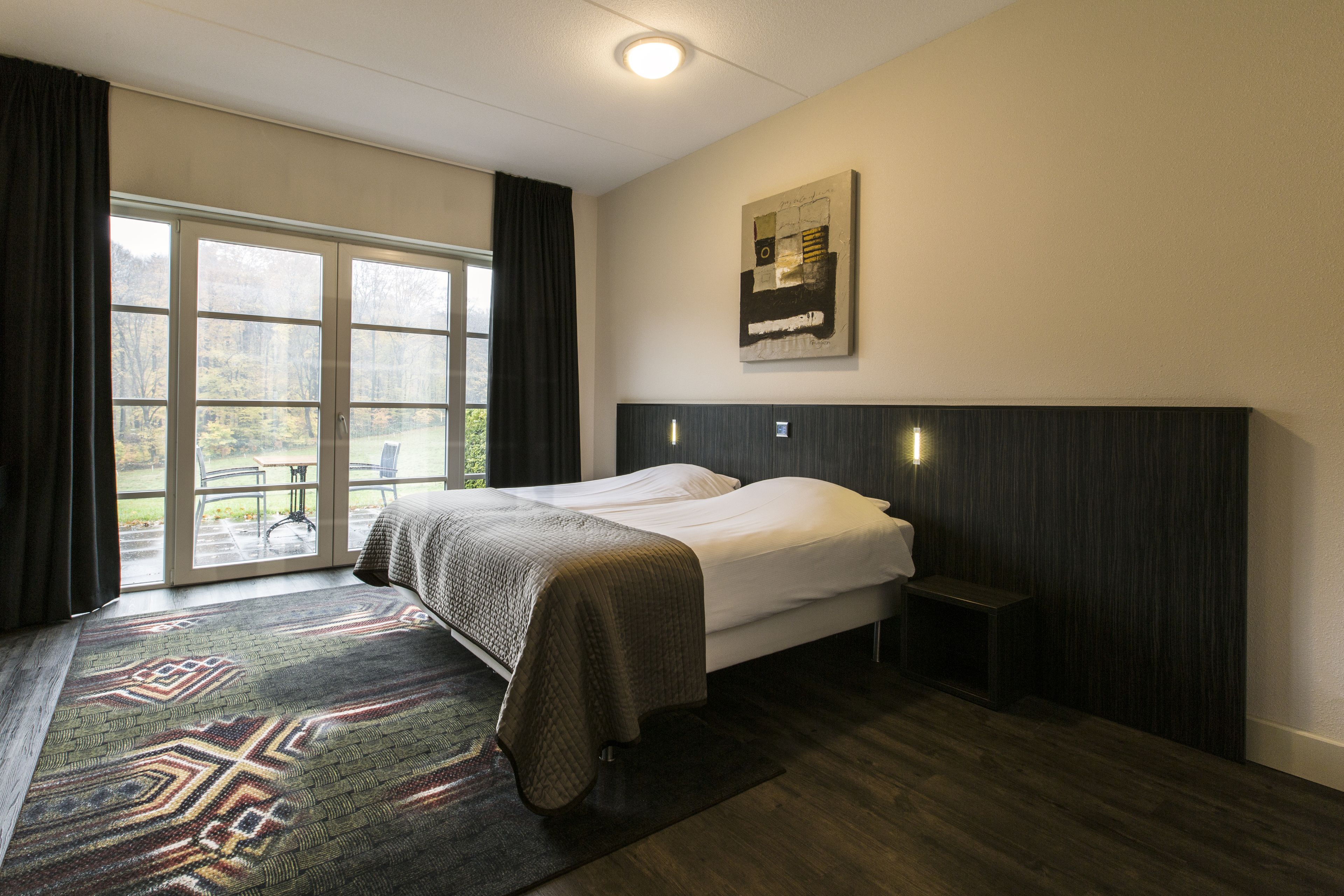 Una habitación de hotel con cama, ventana con cortinas, alfombra y una obra de arte colgada en la pared.