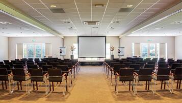 Un salón de conferencias con filas de sillas orientadas hacia una pantalla grande, ventanas laterales y suelo alfombrado.